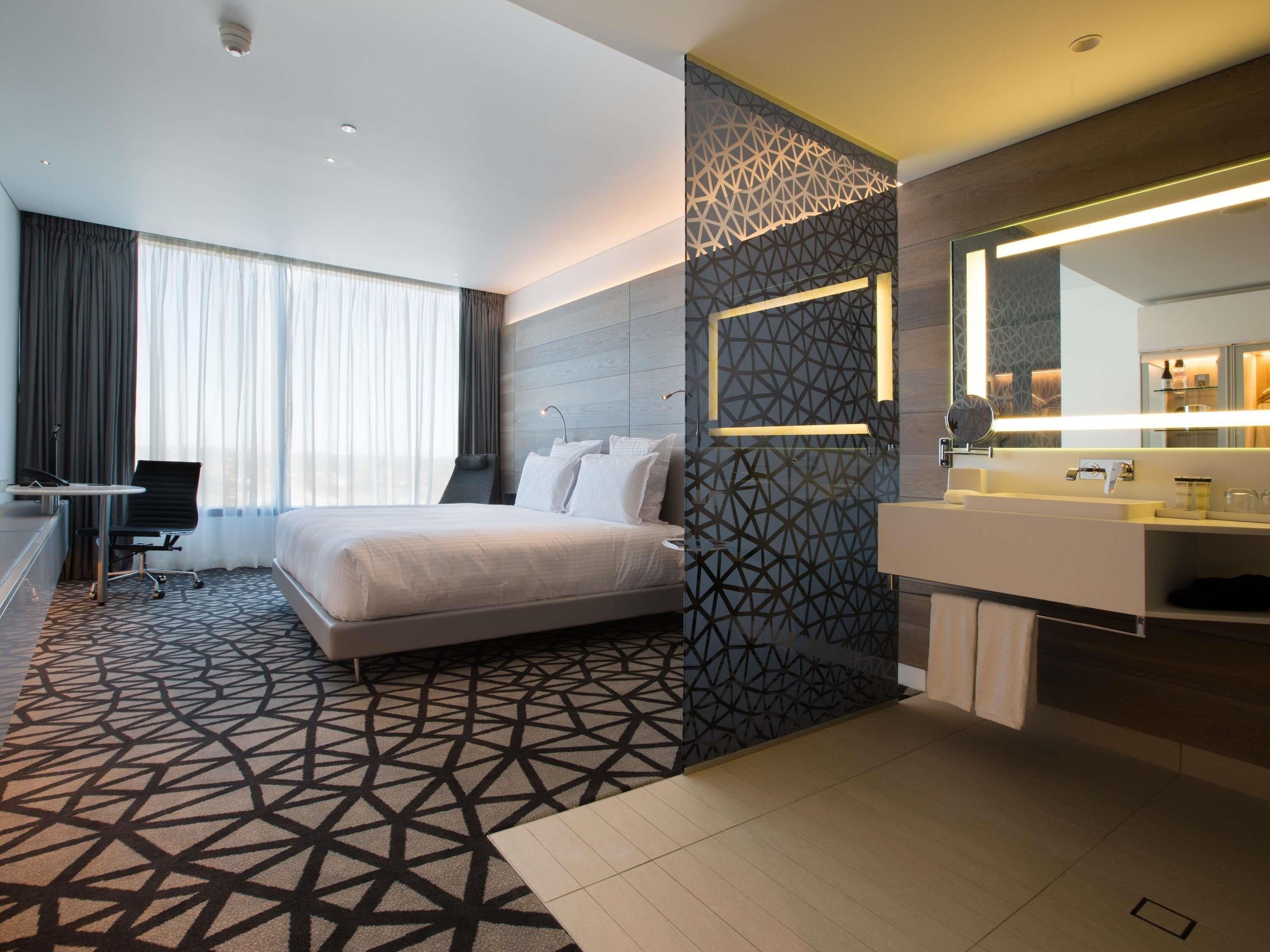 Una habitación de hotel moderna con una cama grande, un escritorio, una silla, un baño con espejo y una pared con estampado geométrico.