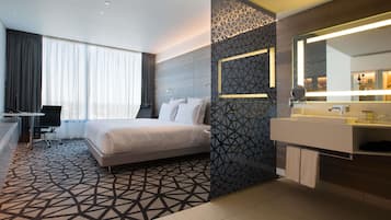 A modern hotel room with a large bed, a desk, a chair, a bathroom with a mirror, and a geometric-patterned wall.