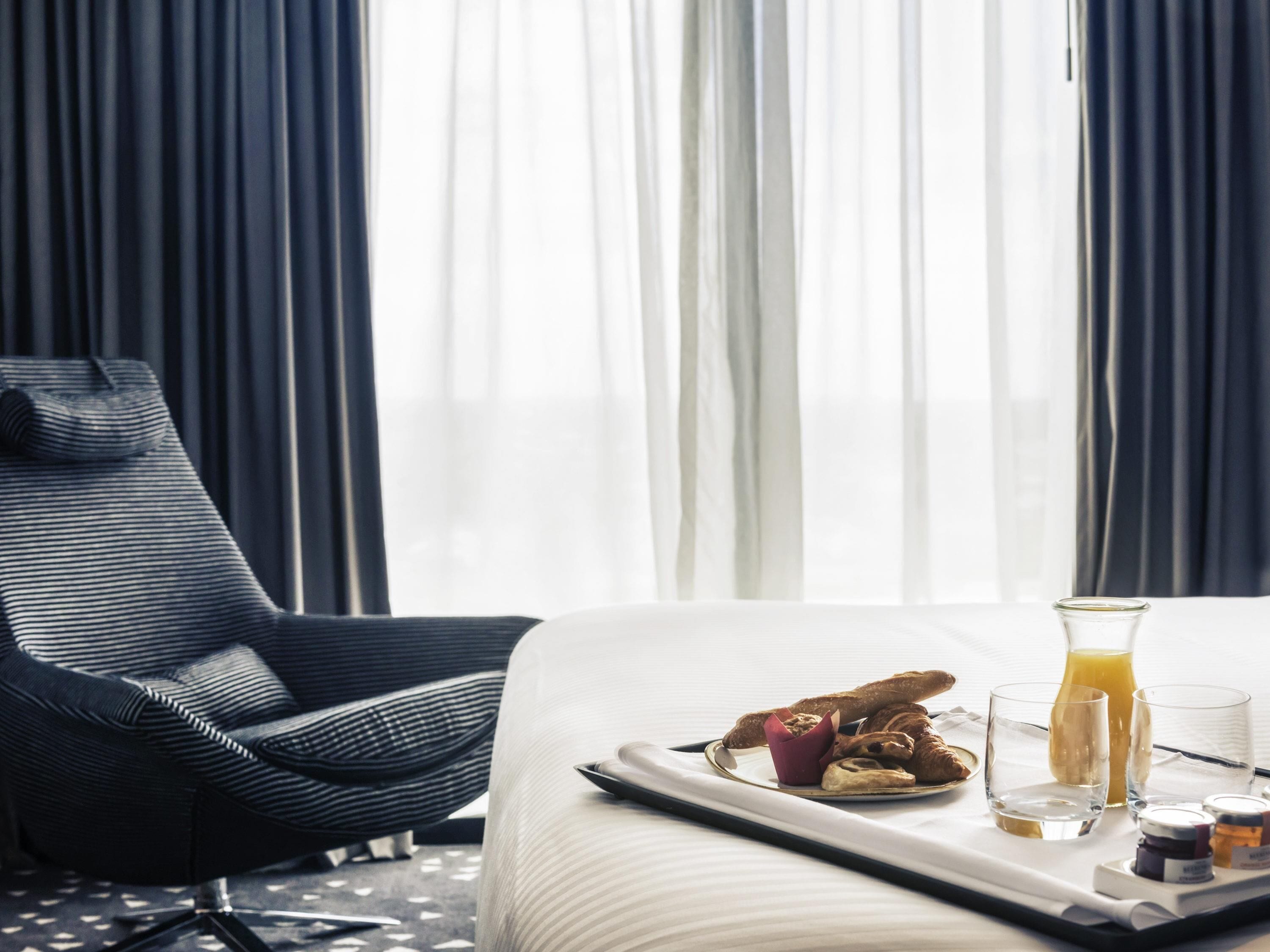 Et hotelværelse med seng, stol, morgenmadstakke og et vindue med gardiner.
