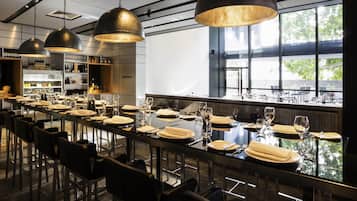 A modern restaurant with a long dining table, black chairs, and pendant lighting.
