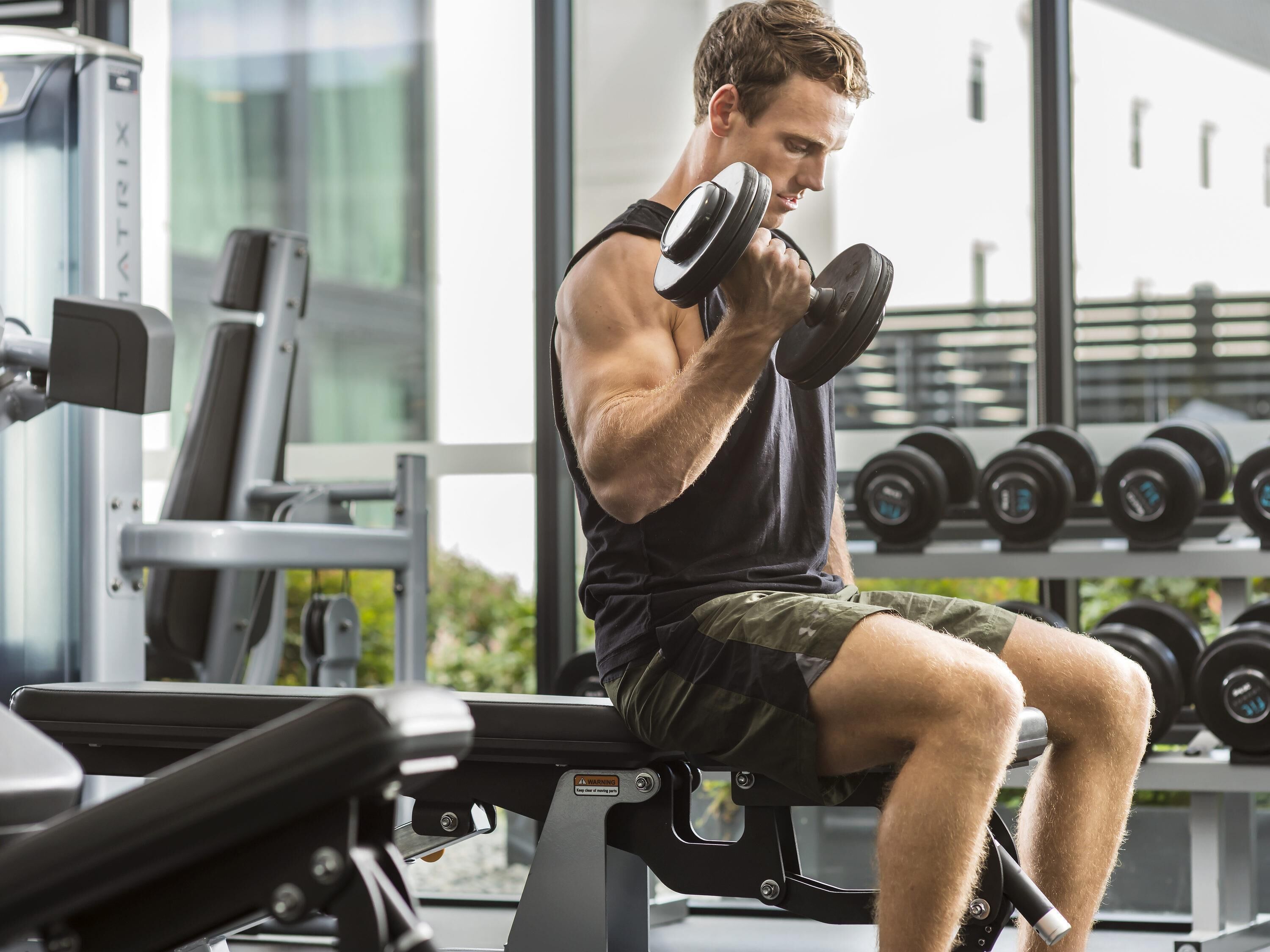 A person exercising with dumbbells in a gym with weight machines and a rack of dumbbells in the background.