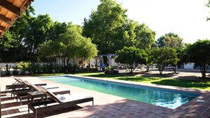 A rectangular swimming pool with a tiled deck, surrounded by trees and a covered area with a pergola.