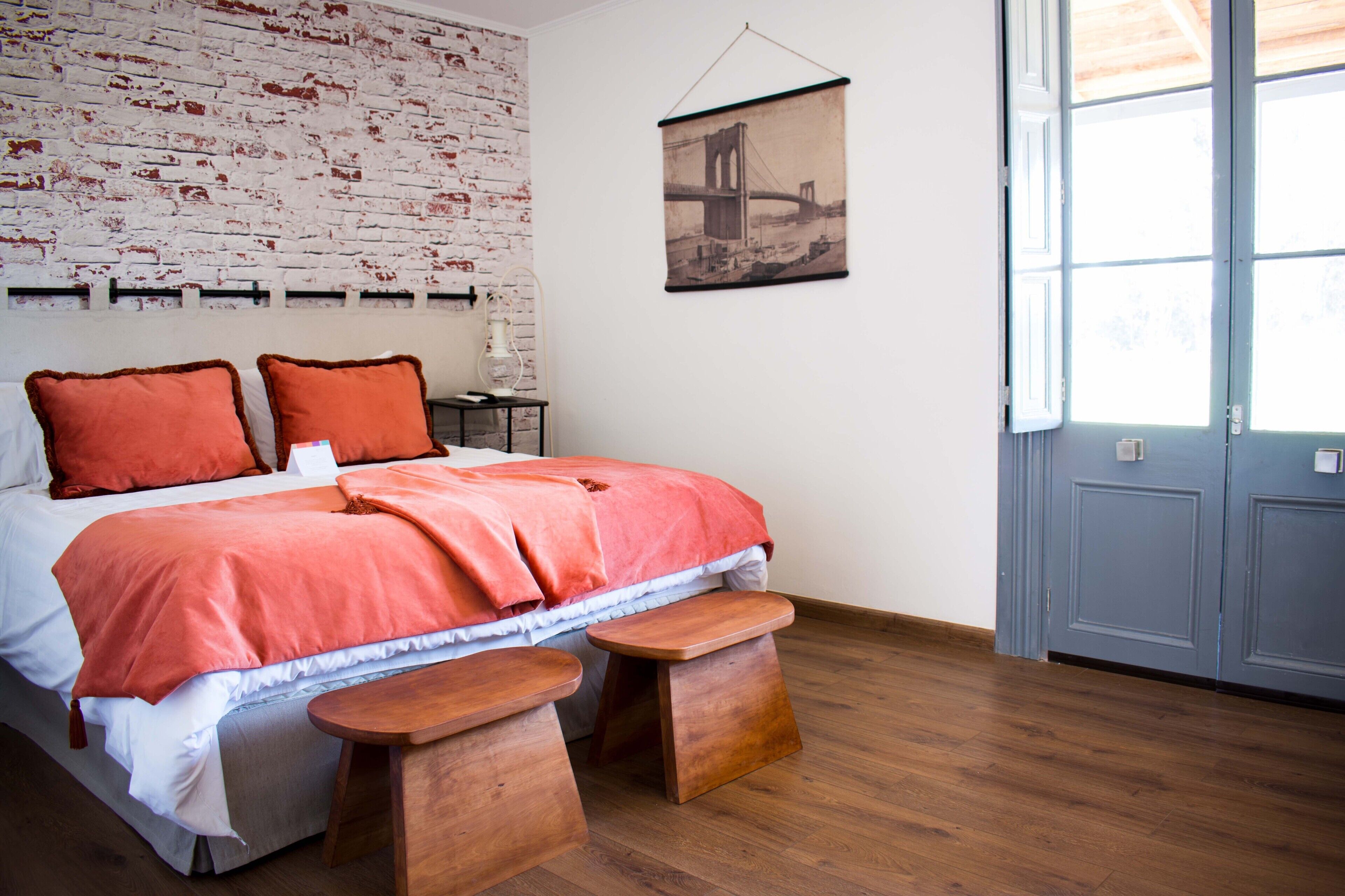 A bedroom with a bed, two pillows, a wooden bench, a small table, a brick-patterned wall, a framed picture, and a door with a window.