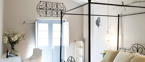 Un lit à baldaquin avec une structure noire, une tête de lit blanche rembourrée et un pied de lit assorti. On y trouve une chaise blanche avec un coussin, une petite table sur laquelle est posé un vase de fleurs et une lampe sur une table de chevet.