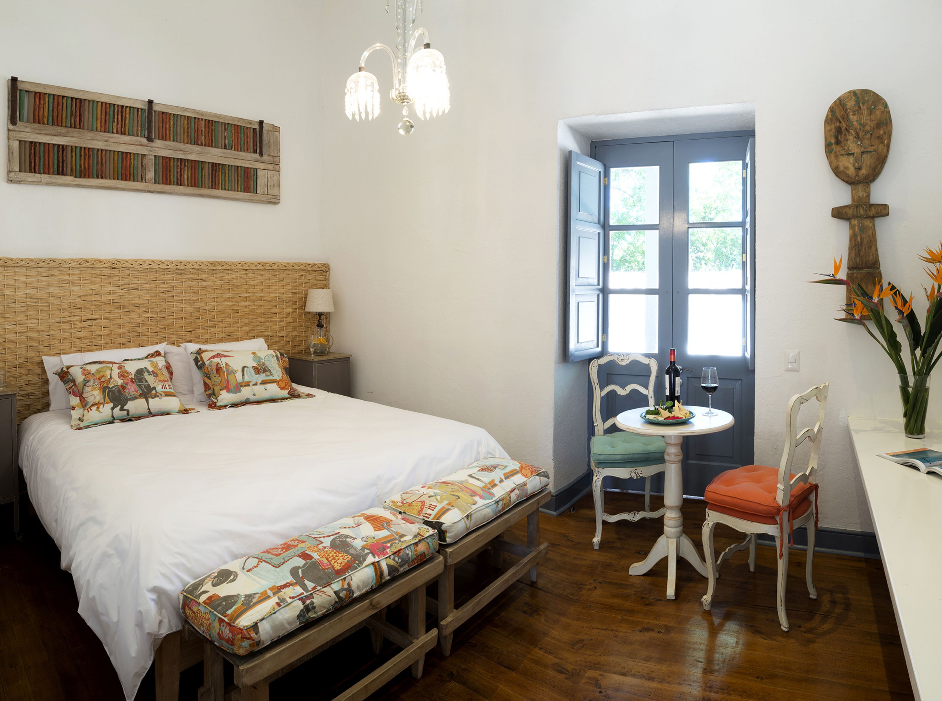Una habitación con una cama, una mesita con una silla, un ventanal y una araña.