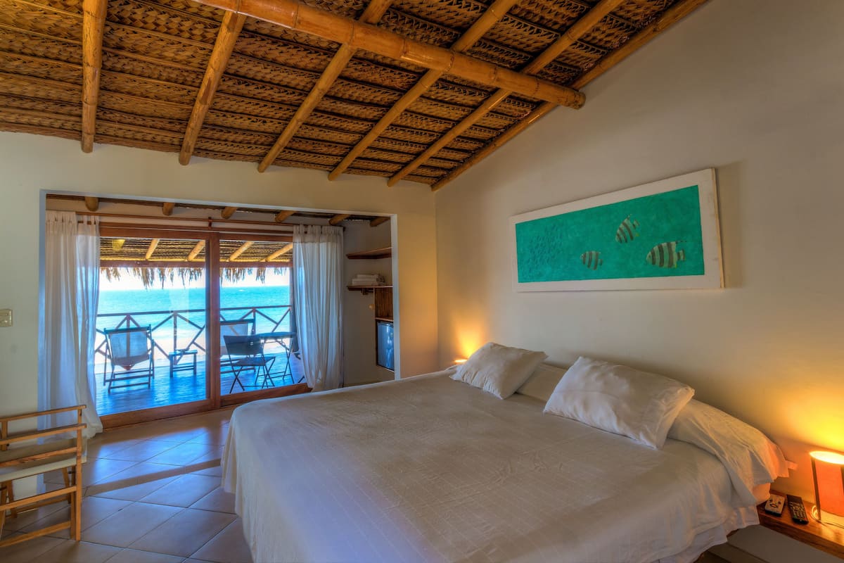 A hotel room with a large bed, a view of the ocean through sliding glass doors, and a wooden ceiling.