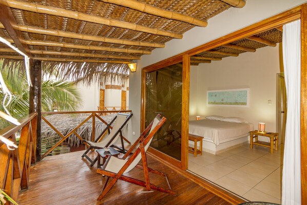 A room with a bed, a small table, and a chair. There is a balcony with a wooden railing and a thatched roof. - Las Olas Mancora (Máncora)