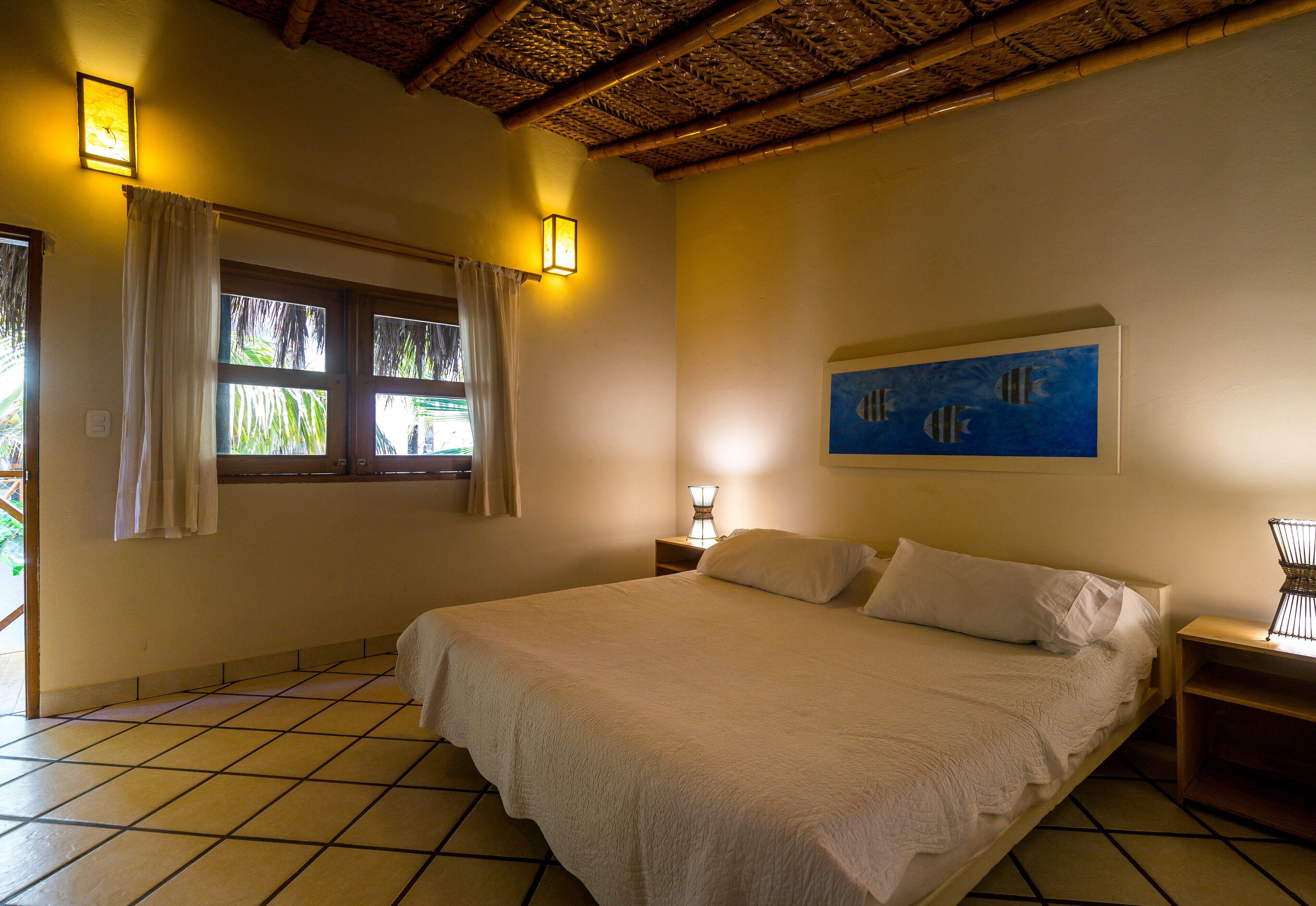 Un dormitorio con cama, mesita de noche, lámpara, cuadro y ventana con cortinas.