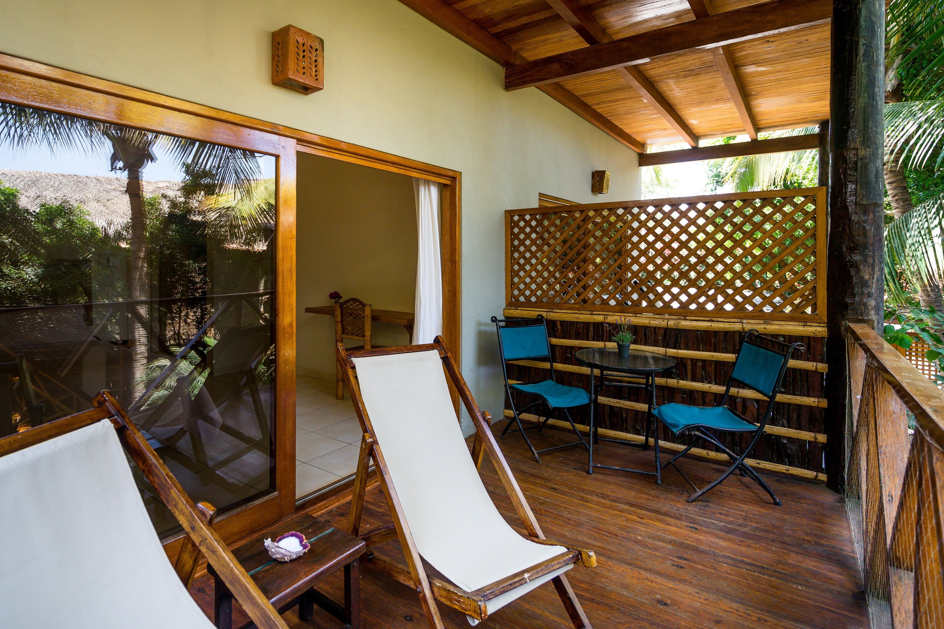Una terraza de madera con un ventanal de vidrio que da a una habitación, una pequeña mesa con sillas y vistas a palmeras.