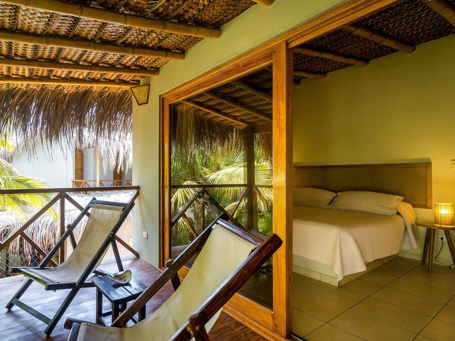 Una habitación con una cama, una mesita de noche, una silla y vistas a palmeras a través de una ventana.