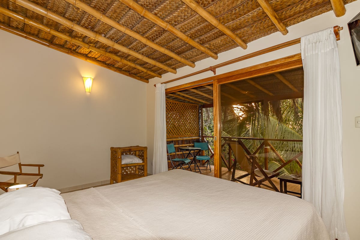 A bedroom with a bed, a nightstand, a chair, and a table. There is a balcony with a view of palm trees.