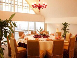 Een eetgedeelte met een ronde tafel, stoelen, een kroonluchter en grote ramen.