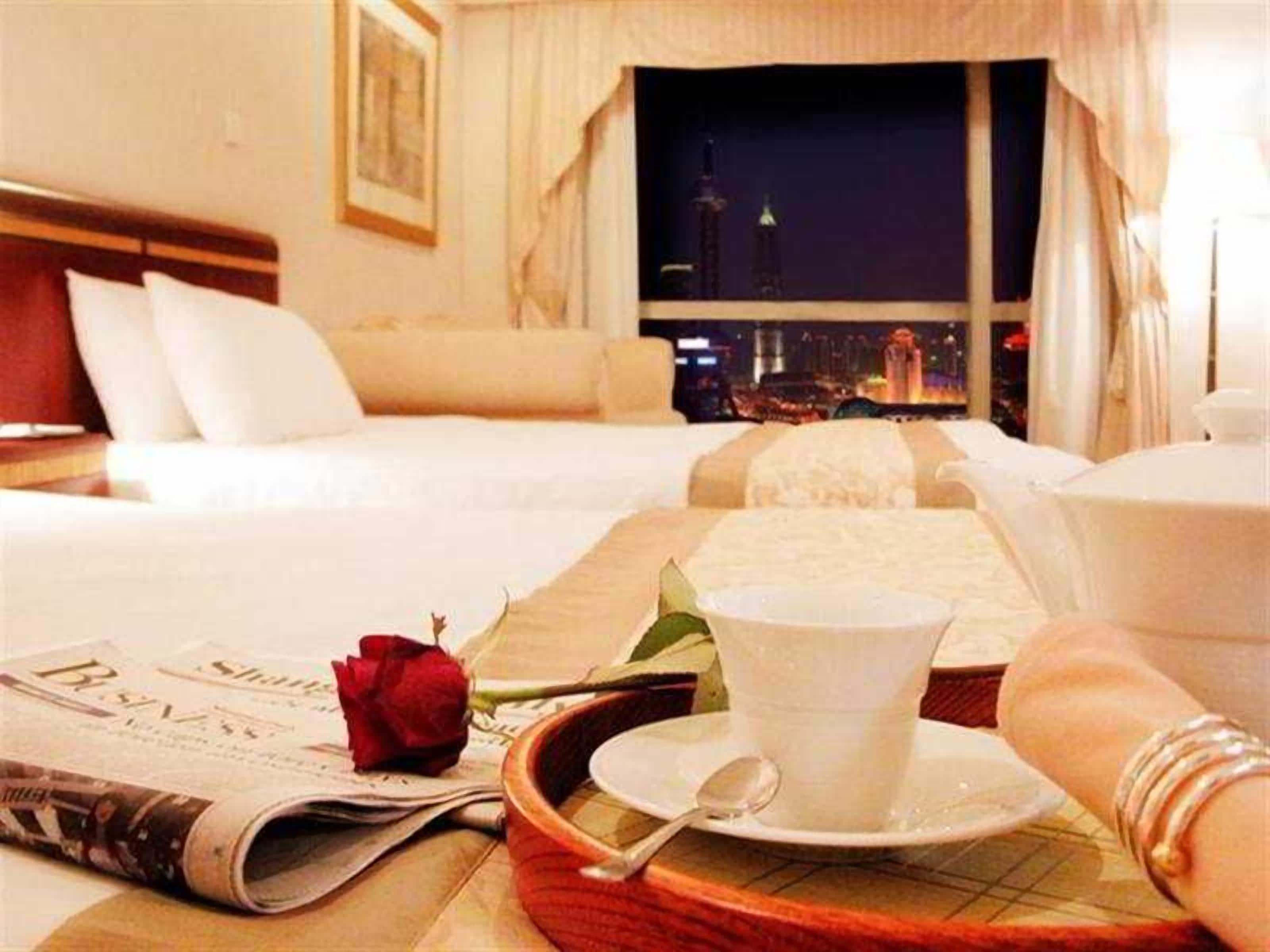 Ein Hotelzimmer mit Bett, Tablett mit Tasse und Untertasse, einer Rose und einer Zeitung.