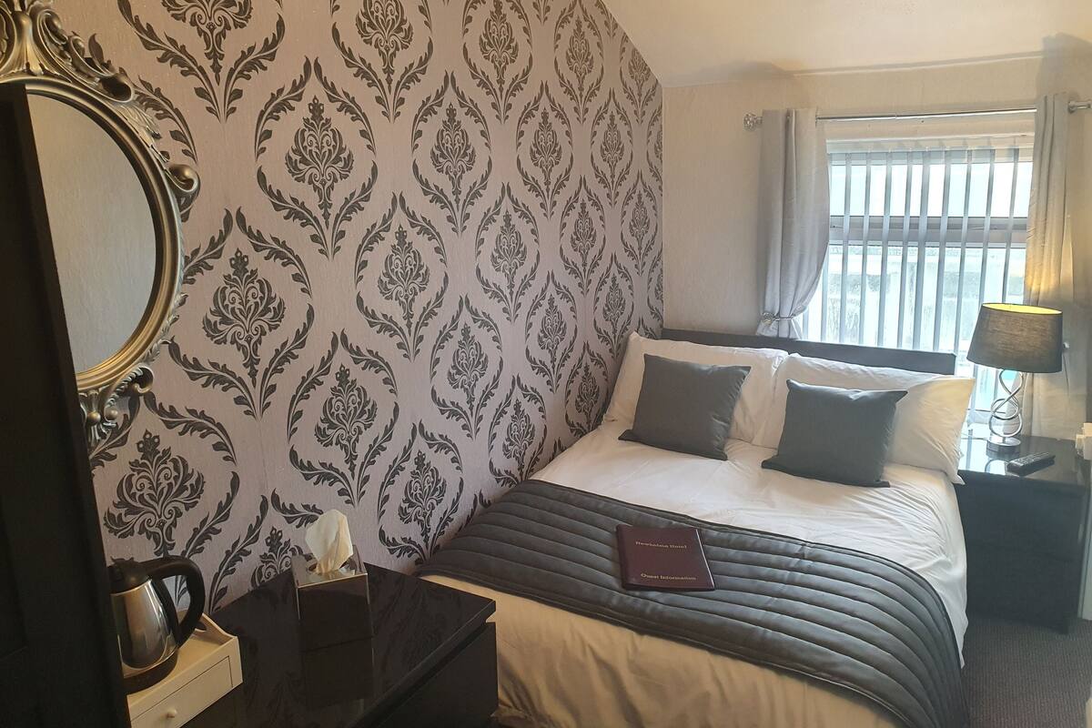 A hotel room with a patterned wallpaper, a bed with pillows, a bedside table with a lamp, and a nameplate on the bed.