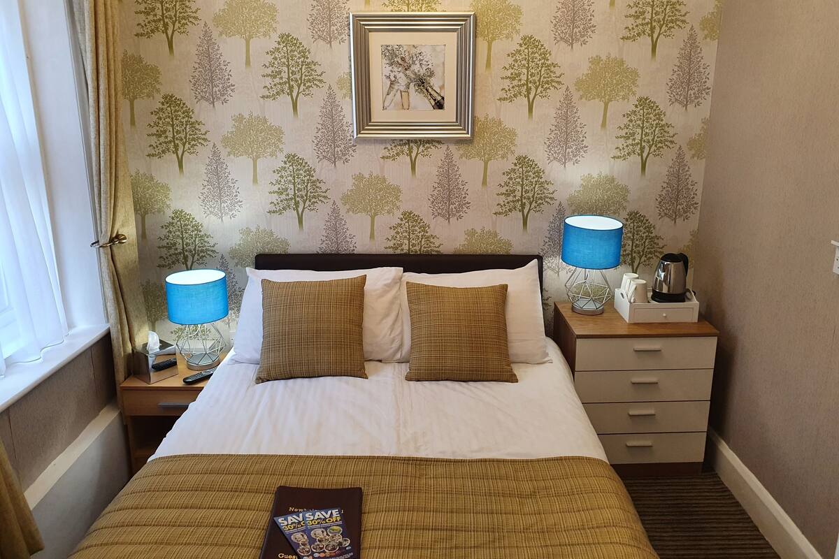 A hotel room with a bed, two bedside tables, a nightstand, a lamp, a framed picture, and a window with curtains.