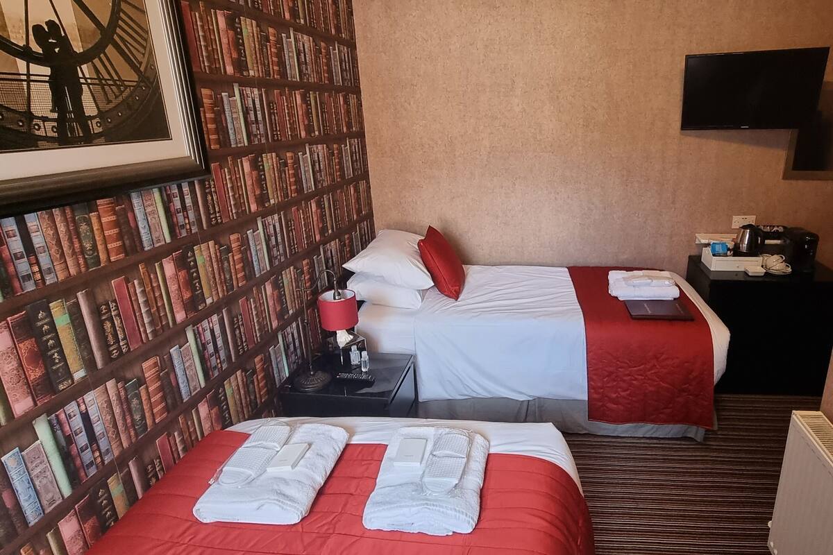 A hotel room with two beds, a bookshelf, a television, and a small table with a kettle and a cup.