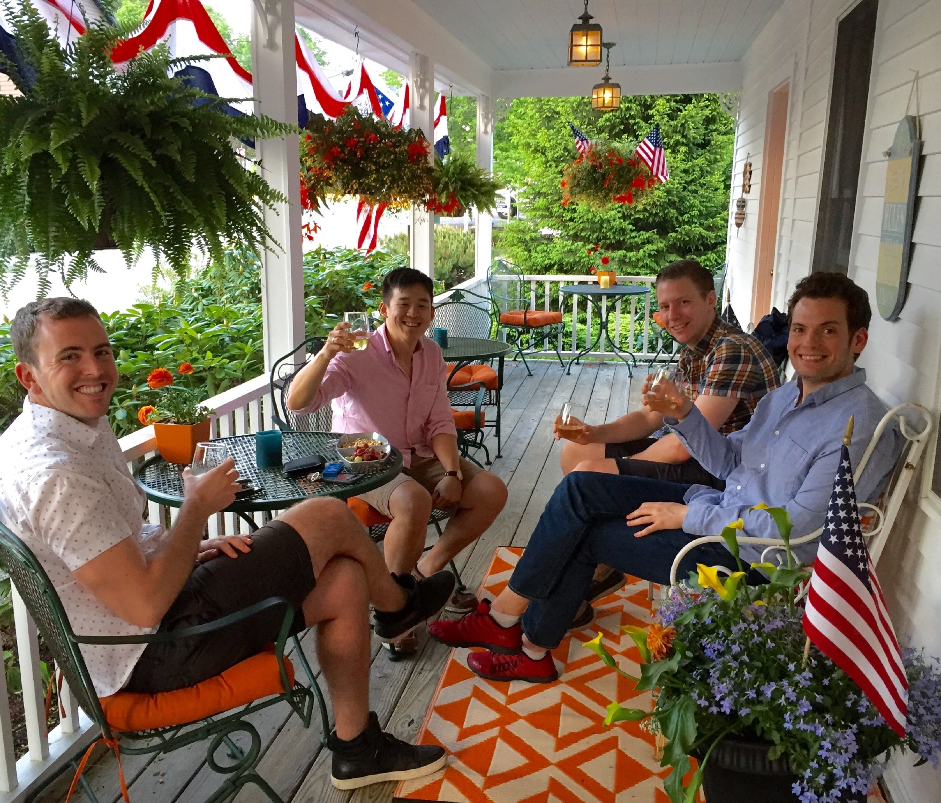 A porch with four people seated on chairs, a table with drinks, and American flags hanging.