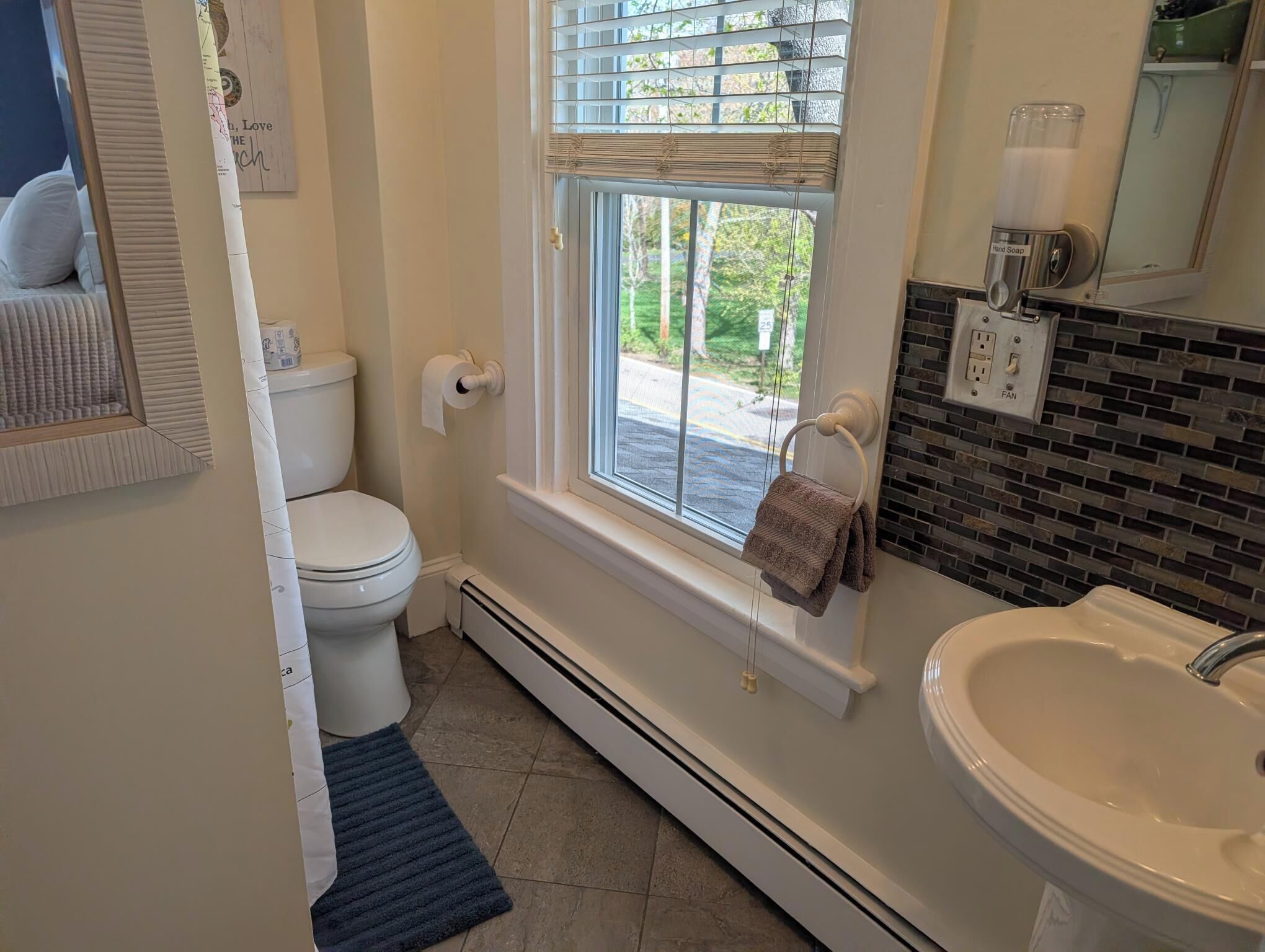 A bathroom with a toilet, a window with a towel rack, a sink, and a hand soap dispenser.