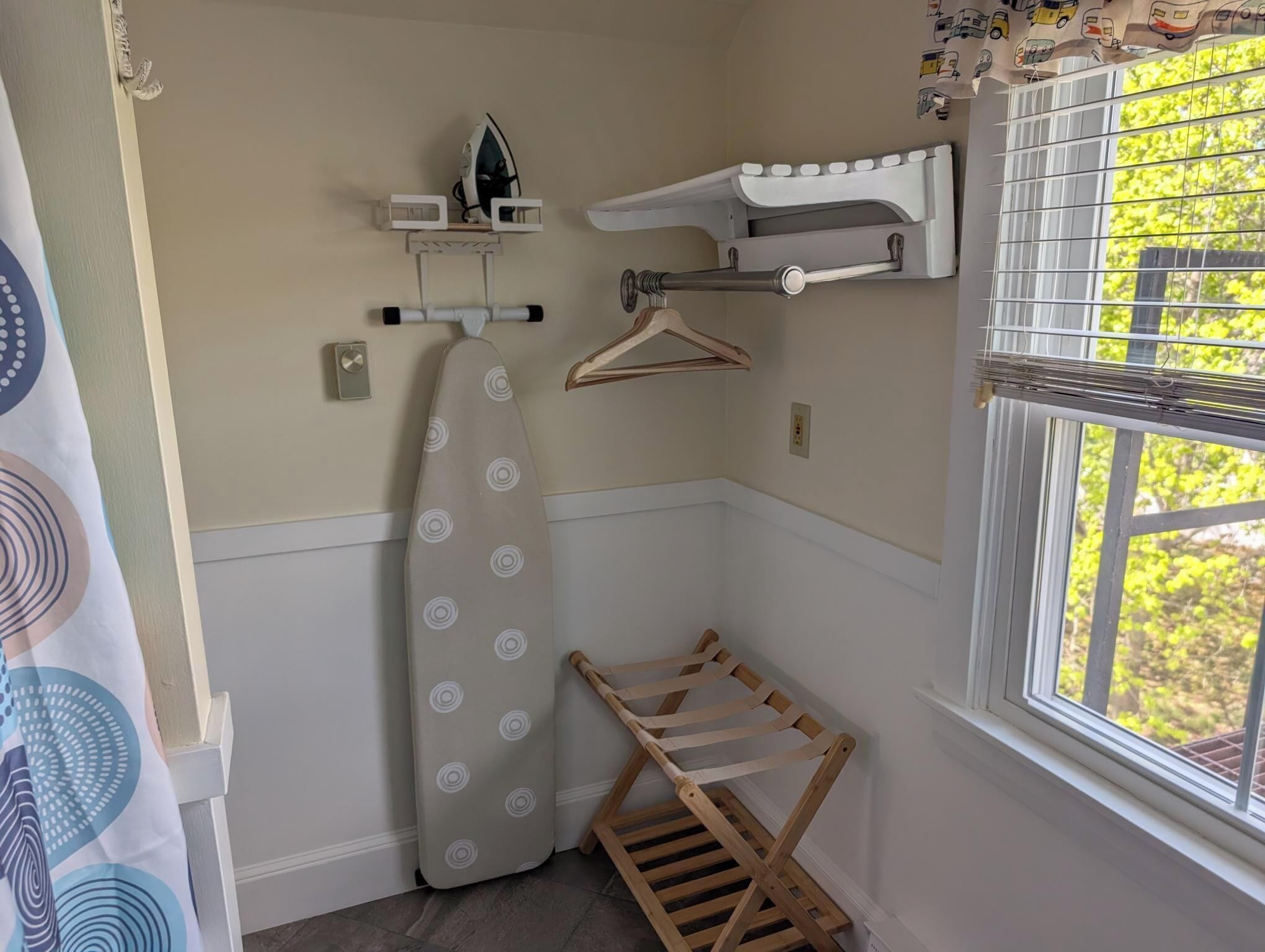 A corner of a room with a hanging iron, a wooden drying rack, and a window with a view of greenery.