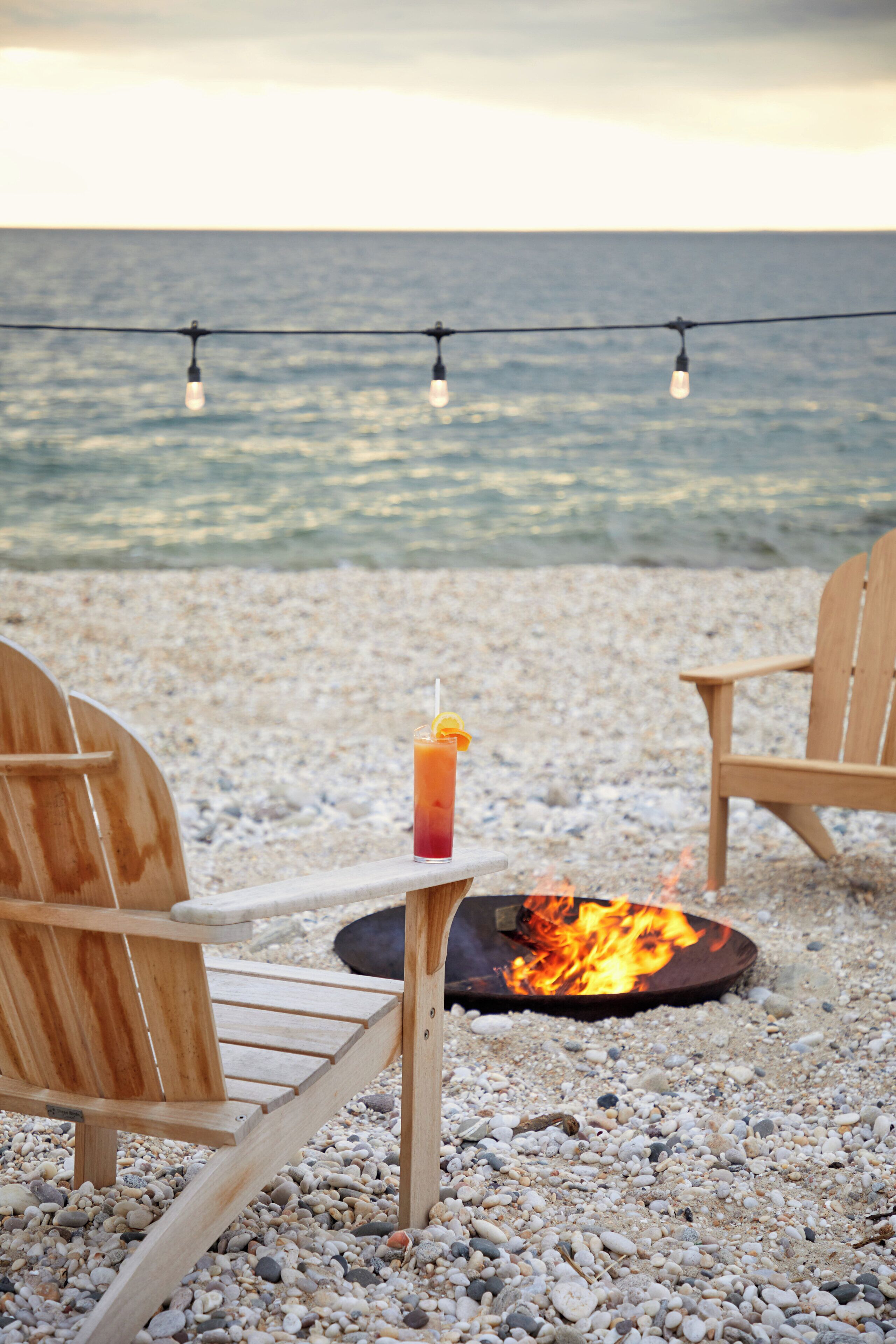 Een strandsetting met twee houten stoelen, een vuurkuil en een drankje op een tafel.