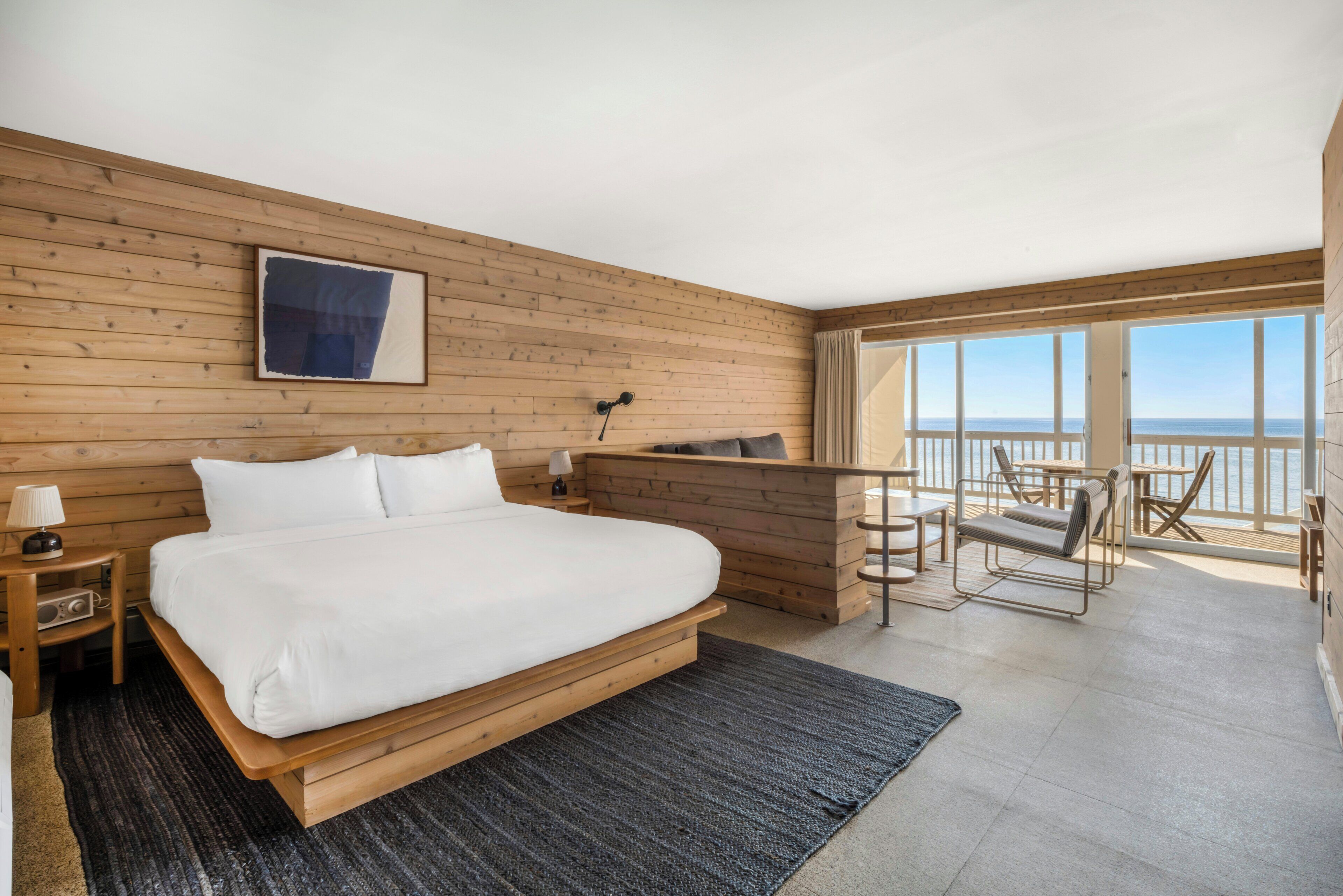 A bedroom with a large bed, wooden panel walls, a desk, and a balcony with a view.