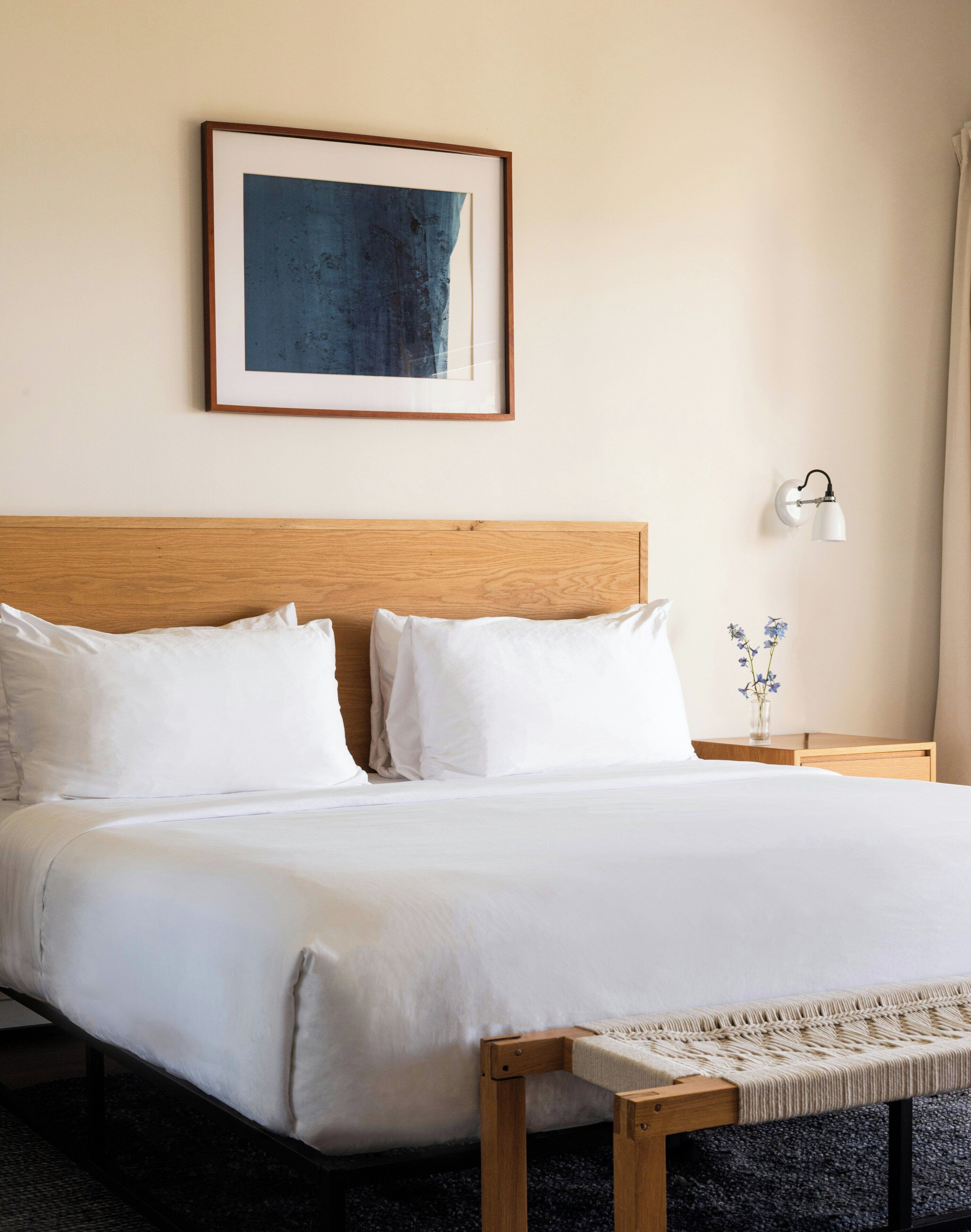 A neatly made bed with white linens, a wooden headboard, a framed abstract painting, a bedside lamp, and a small vase with flowers.