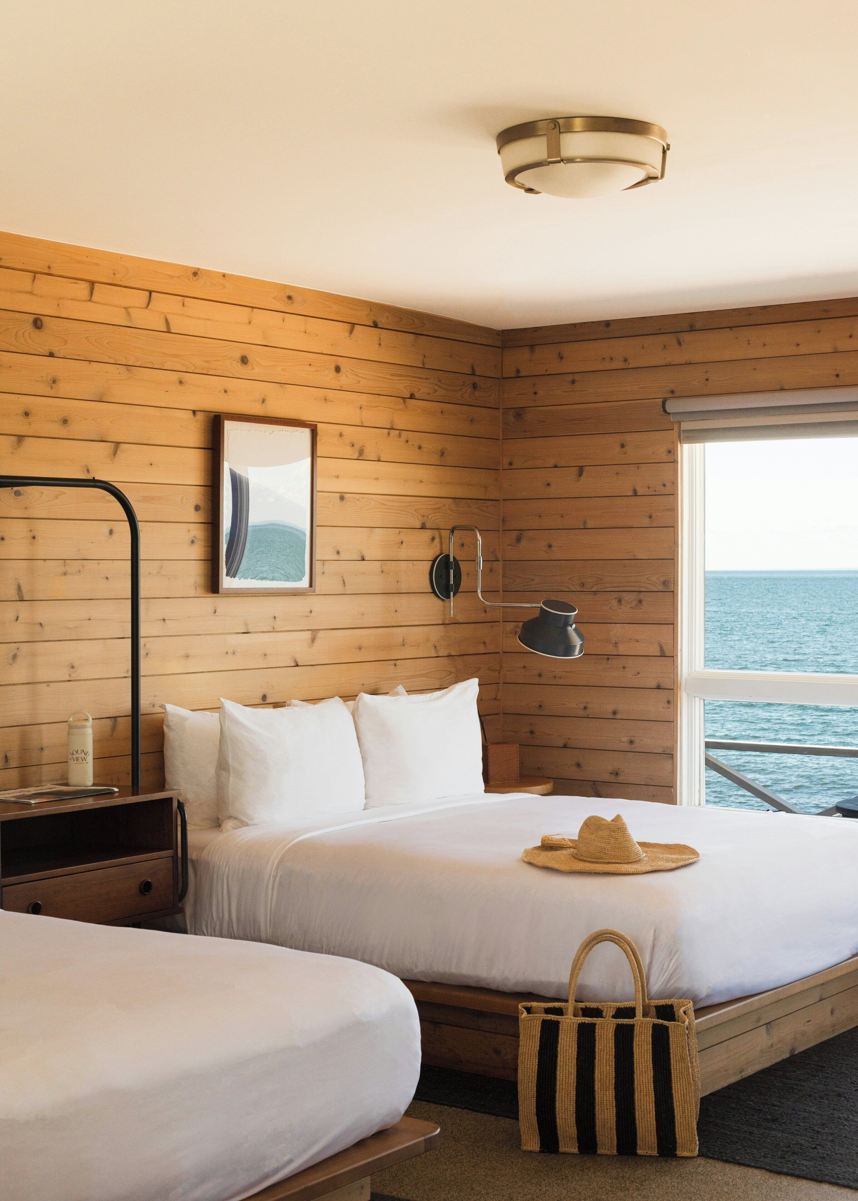 Habitación con paneles de madera, cama con ropa blanca, mesita de noche con lámpara, un cuadro en la pared y vista al mar a través de una ventana.