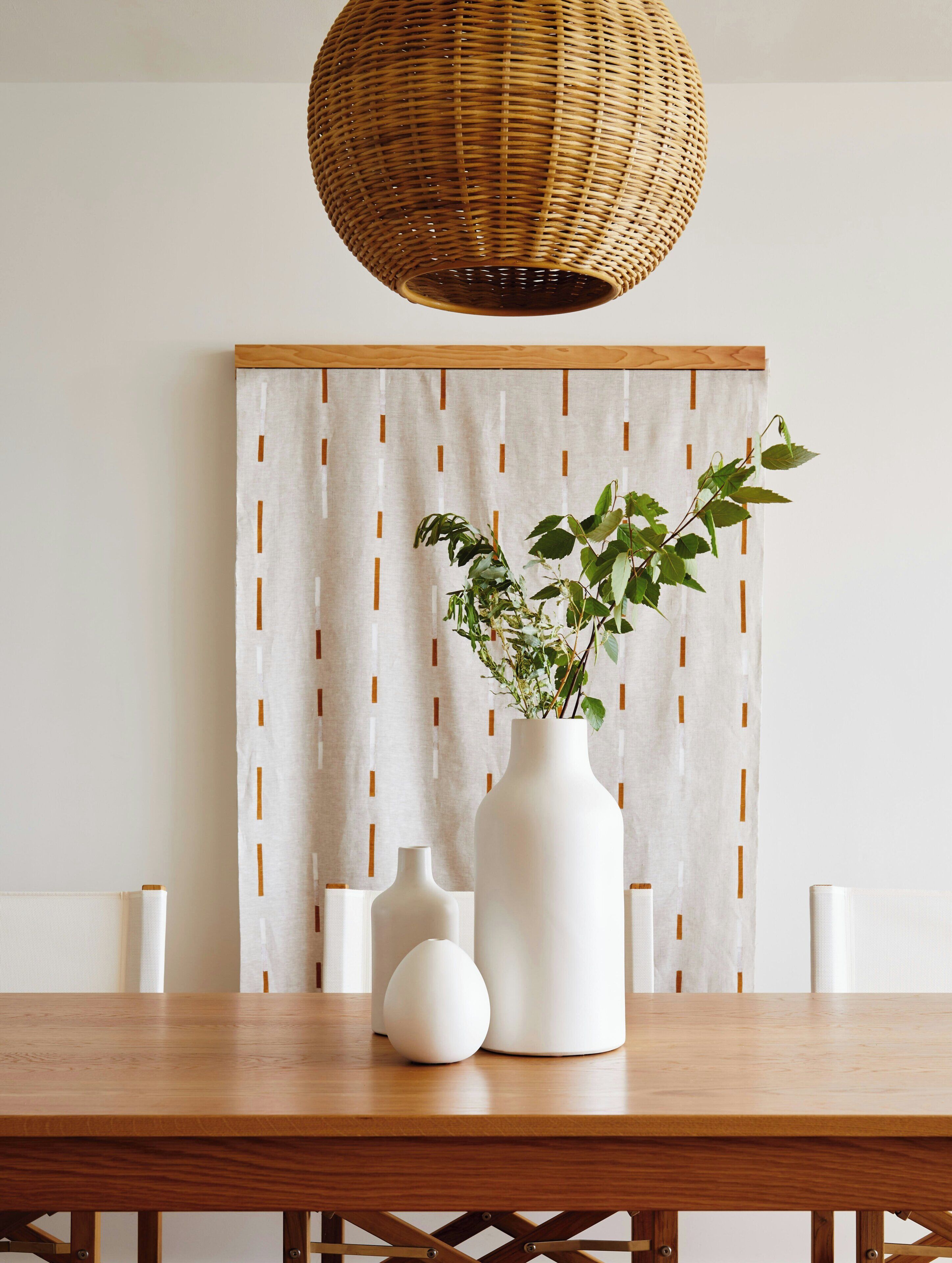 Una mesa de comedor con un jarrón blanco y velas, una lámpara colgante de fibra natural y un fondo decorado.