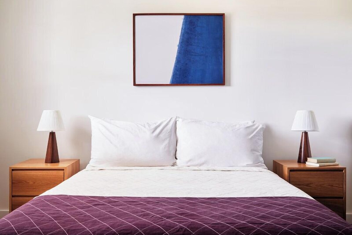 A hotel room with a bed, two bedside tables, a nightstand, a purple bedspread, and a blue and white abstract painting on the wall.