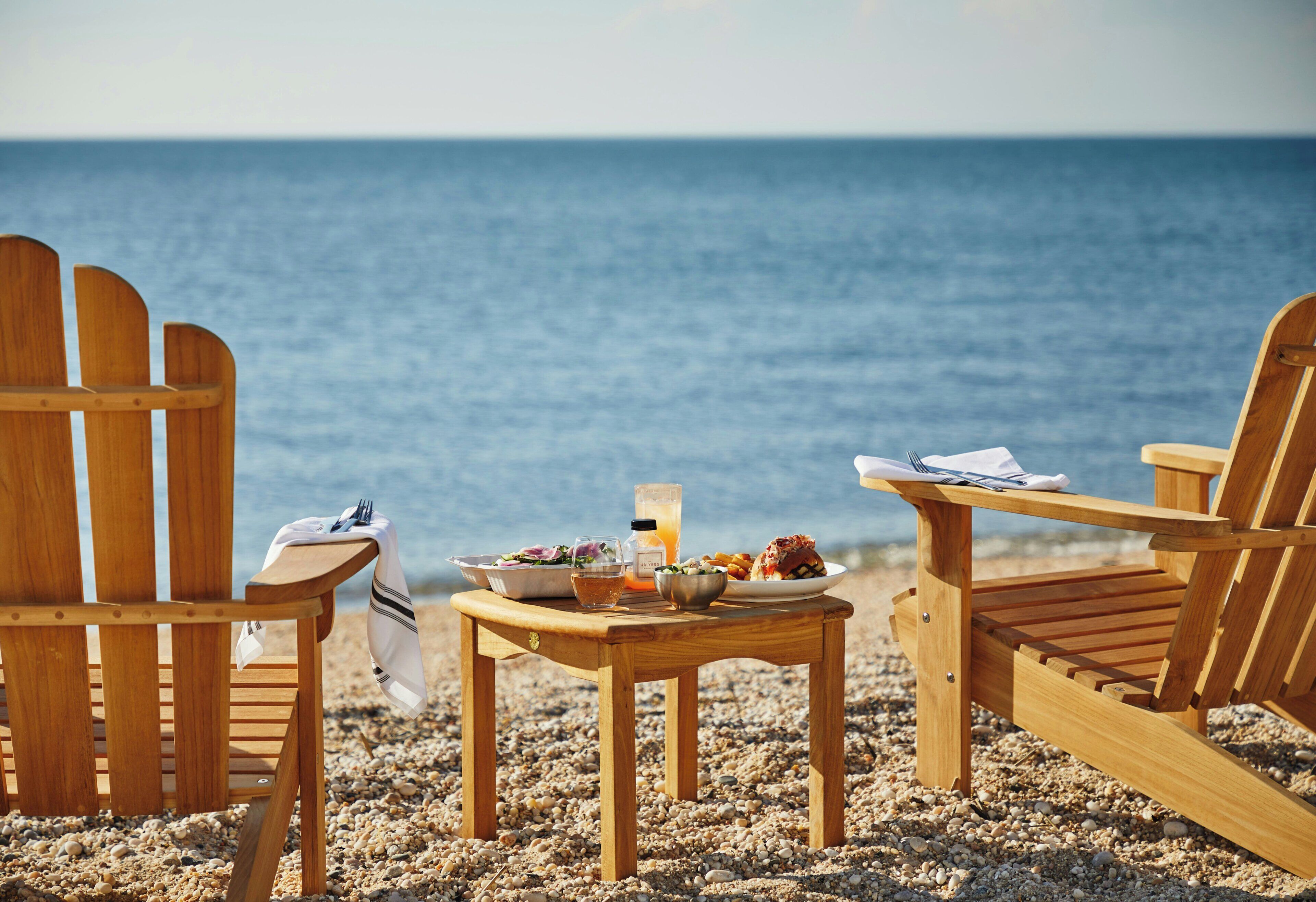 Zwei hölzerne Adirondack-Stühle stehen auf einem Sandstrand, dazwischen ein Tisch mit Essen und Getränken.