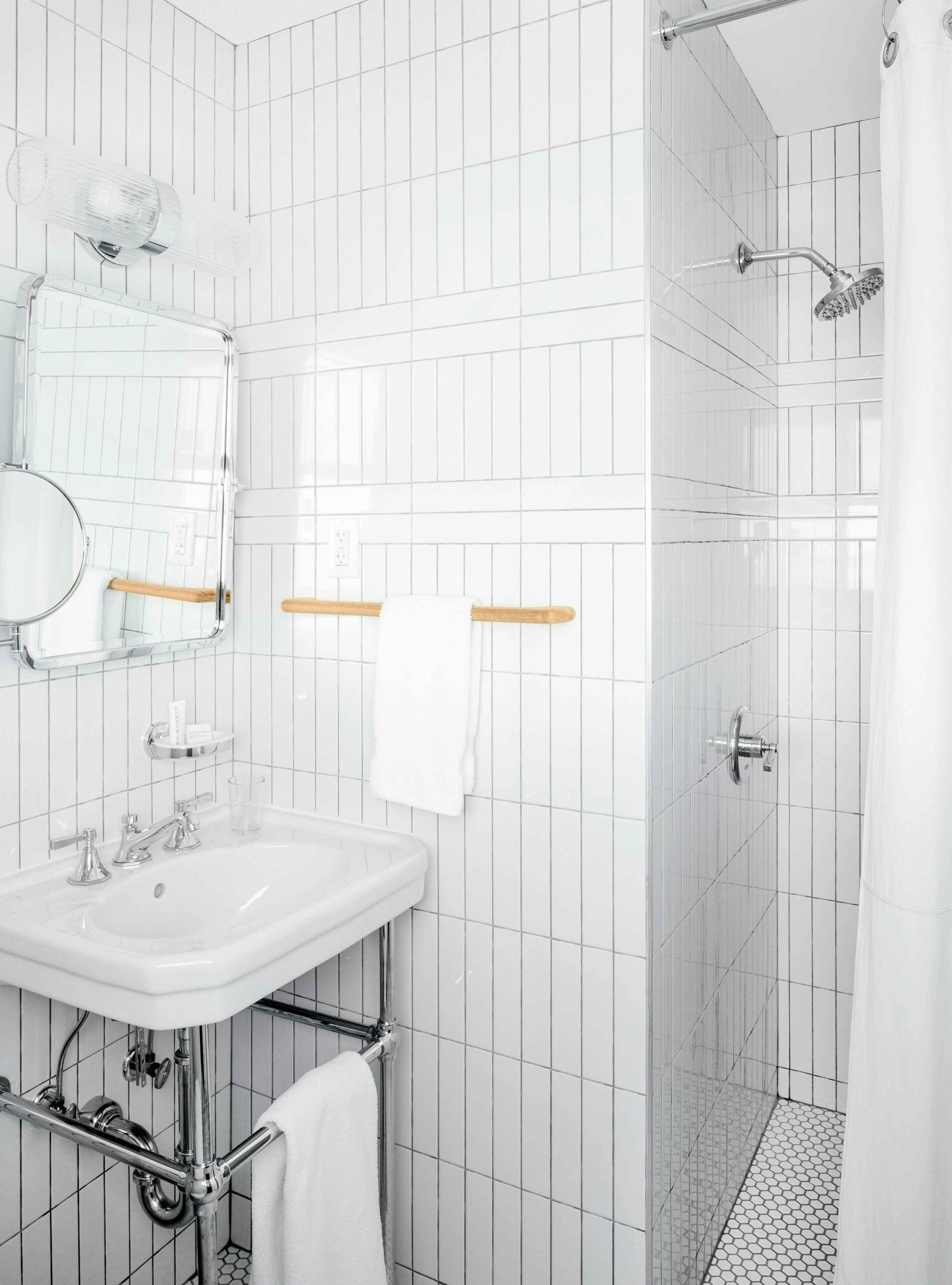 A modern bathroom with white tiles, a sink, a mirror, a towel rack, and a shower.