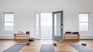 A spacious room with large windows, a wooden bench, yoga mats, and a door leading to a balcony with a view of the sea. - Sound View Greenport (Greenport)