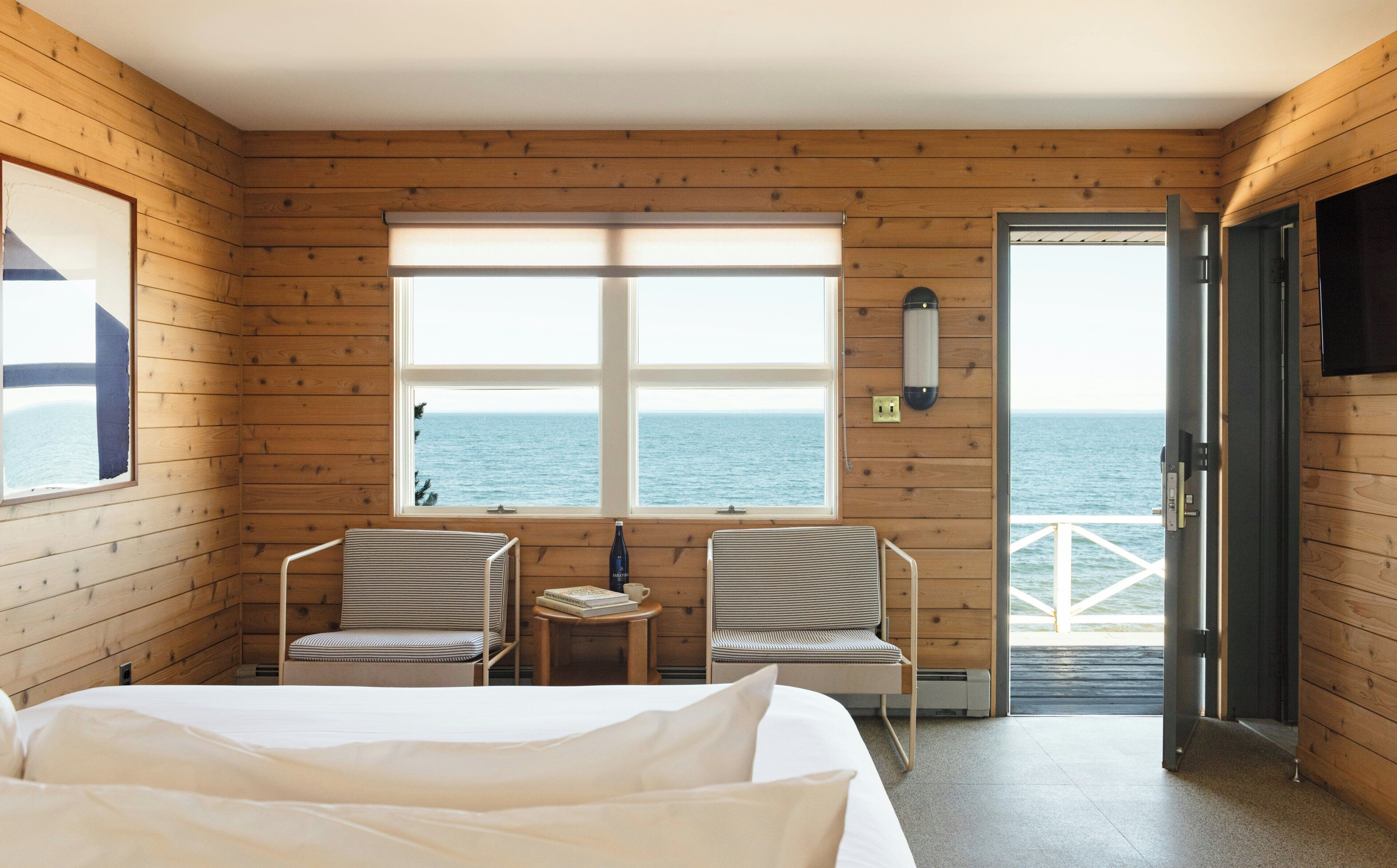 Una habitación con una cama, dos sillas a rayas, una mesita con una botella y un libro, un ventanal con vista al mar y una puerta que da al exterior.