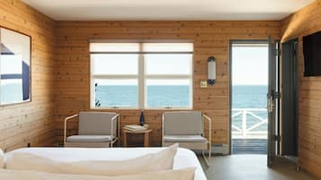 A room with a bed, two striped chairs, a small table with a bottle and a book, a window with a view of the sea, and a door leading outside.