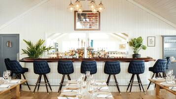 Interior de restaurante con techo alto, zona de bar con gran variedad de botellas y disposición de mesas y sillas.