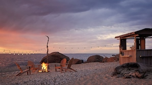 A beach scene with a wooden structure, two wooden chairs, a campfire, and string lights. - Sound View Greenport (Greenport)