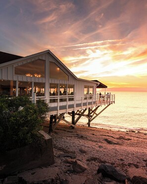A coastal building with a deck extending over the water, featuring large windows and a dining area.