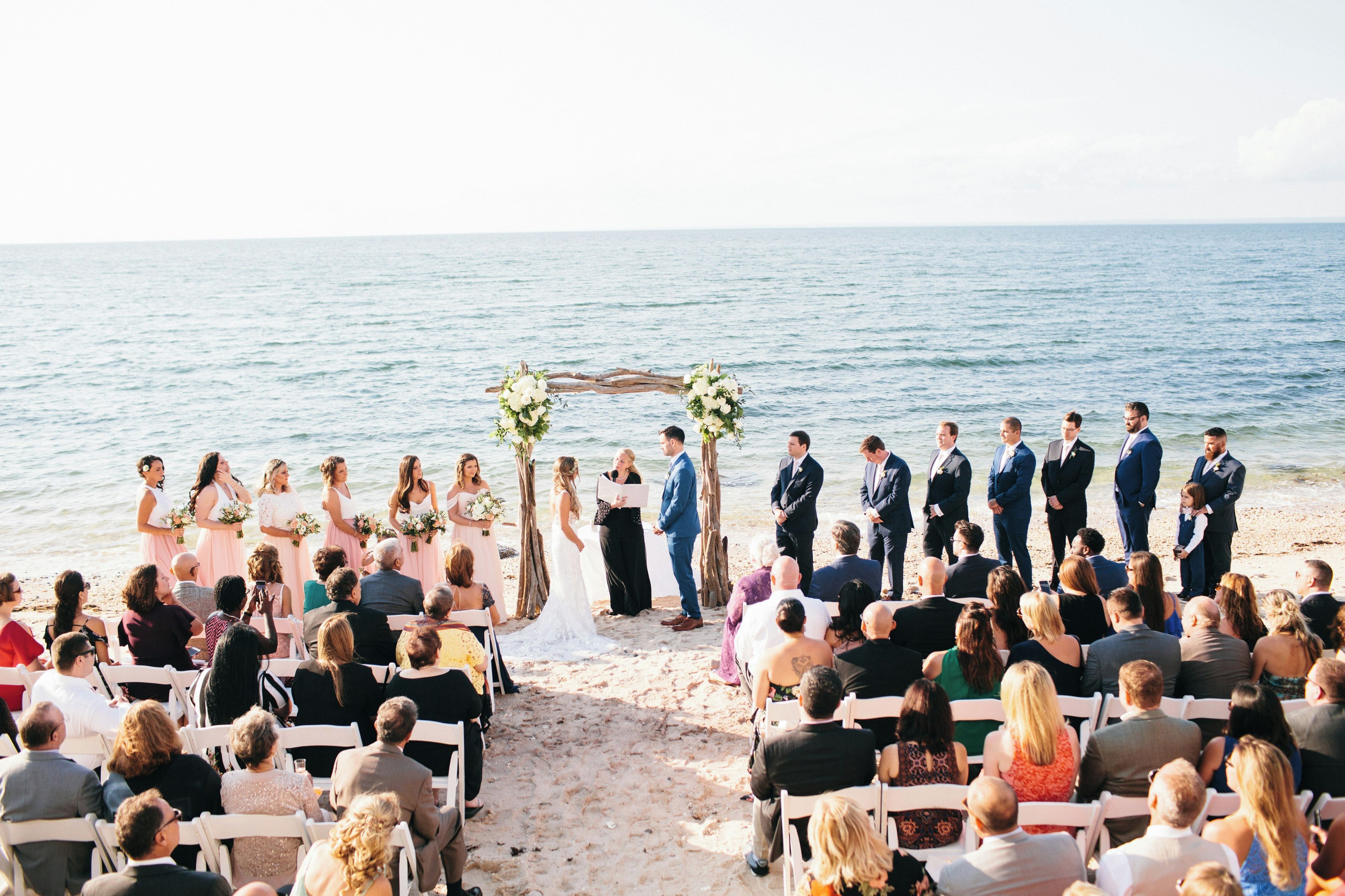 Una ceremonia de boda en la playa con invitados sentados en sillas blancas frente al pasillo.