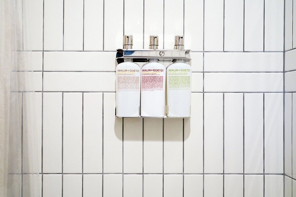A bathroom with white tiled walls and three bottles of shampoo on a shelf.