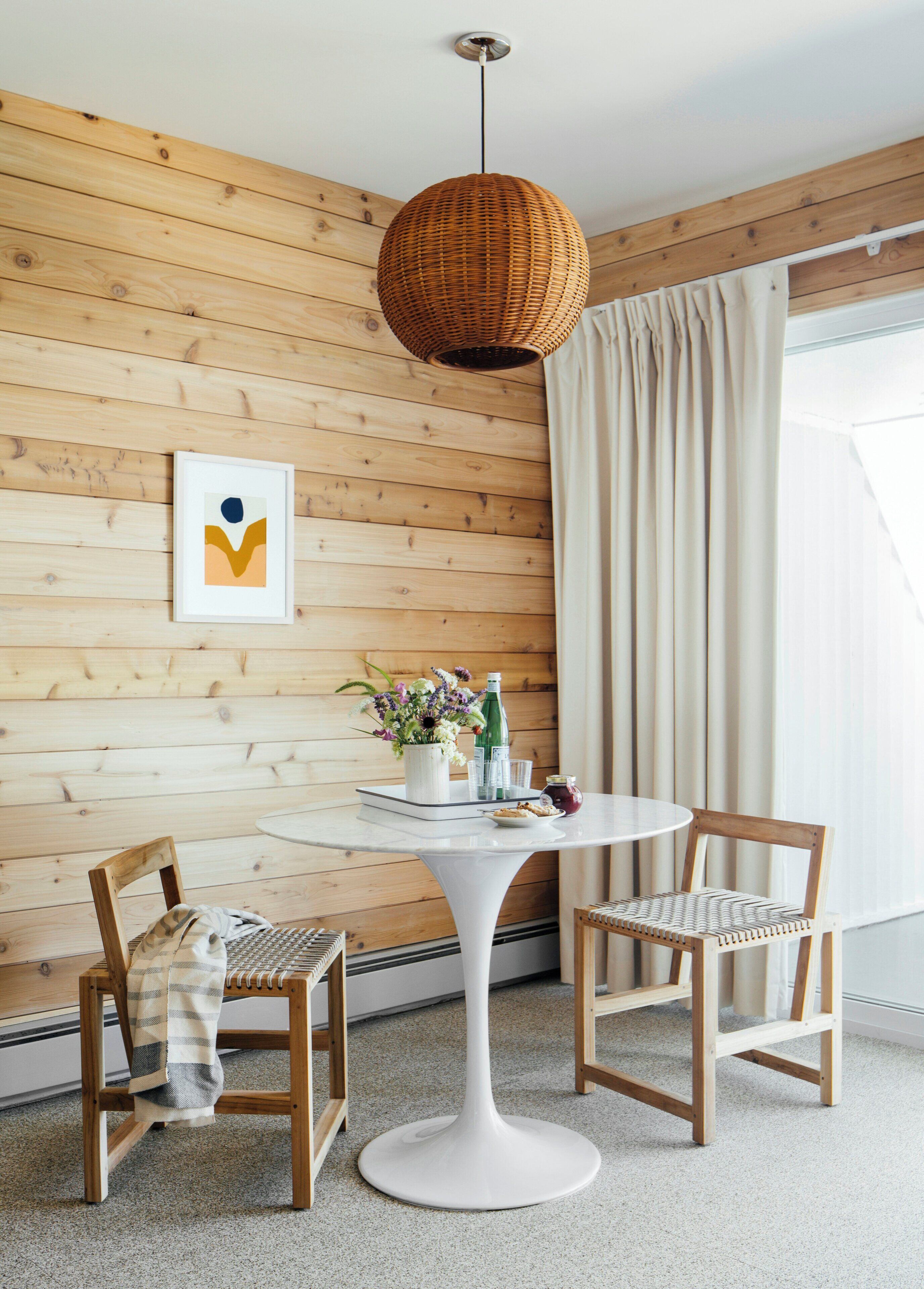 Um quarto com painéis de madeira, uma mesa redonda branca com cadeiras, um candeeiro de vime pendente e um quadro na parede.