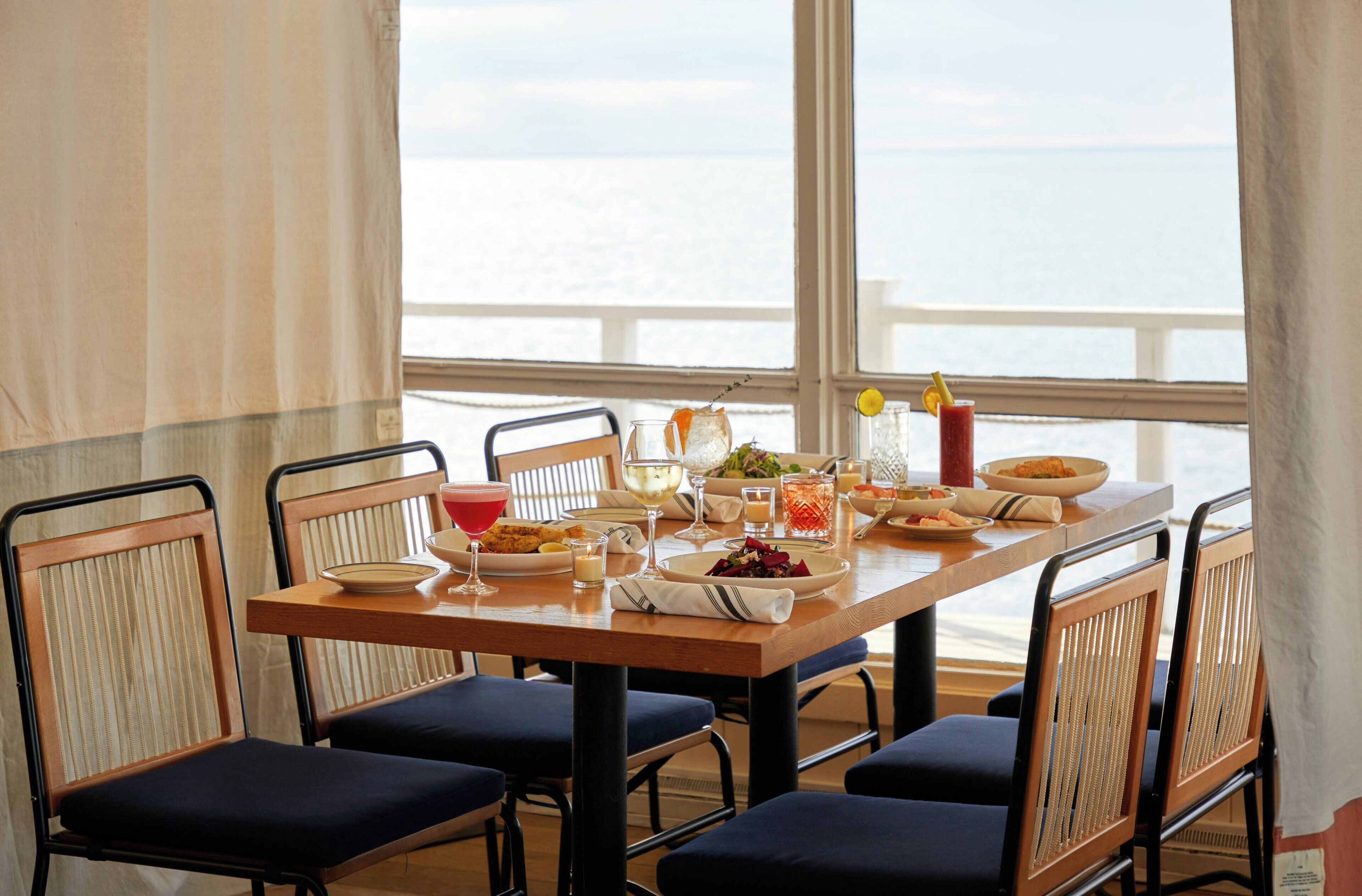 Une table à manger dressée pour quatre personnes, avec vue sur la mer grâce à de grandes fenêtres.