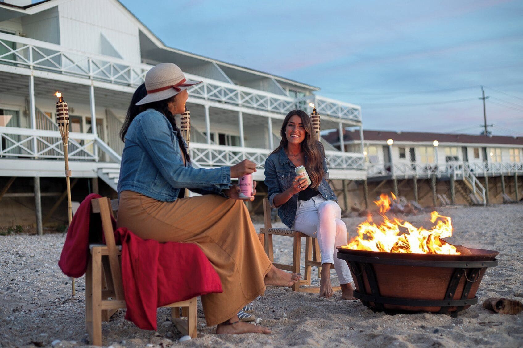Zwei Personen sitzen an einem Lagerfeuer auf einem sandigen Strand, im Hintergrund ein Gebäude.