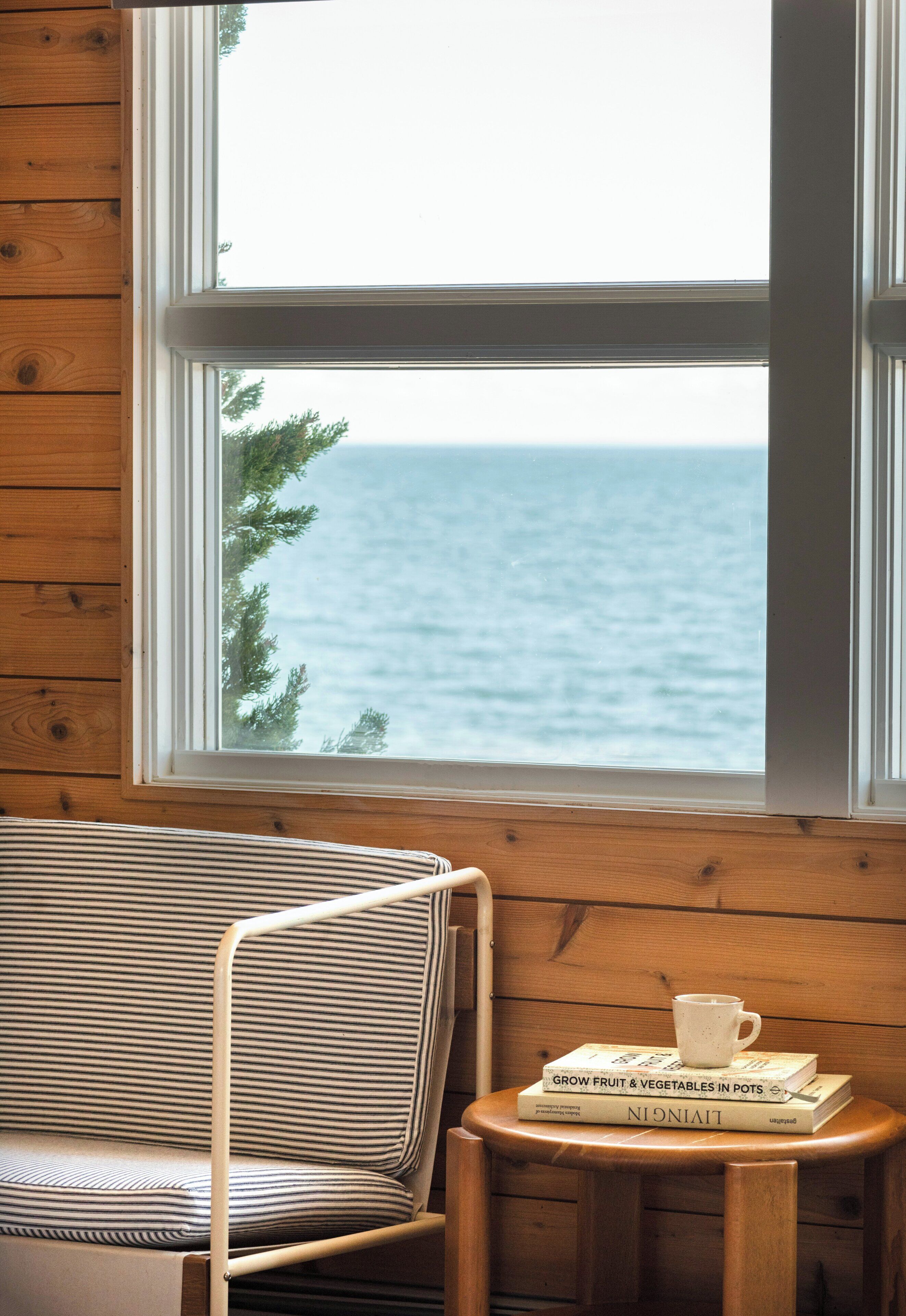 Un interior de madera con vista al mar a través de una ventana, una silla a rayas, y una pequeña mesa con libros y una taza.