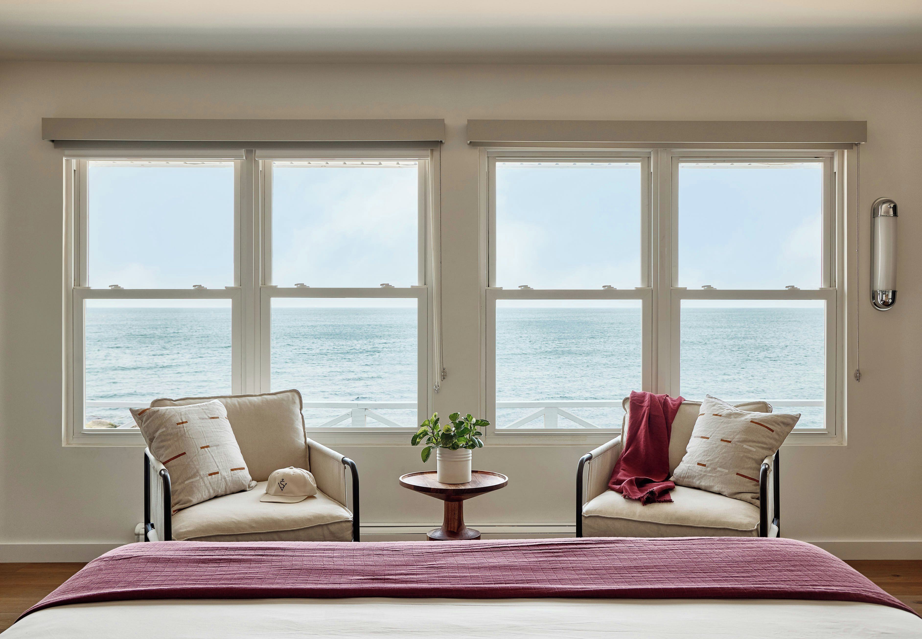 Une chambre avec un lit, deux fauteuils, une petite table avec une plante, et une vue sur la mer grâce à de grandes fenêtres.