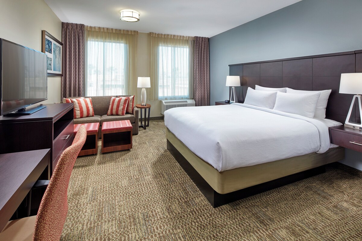 A hotel room with a large bed, a desk with a TV, a sofa, two side tables with lamps, and a carpeted floor.