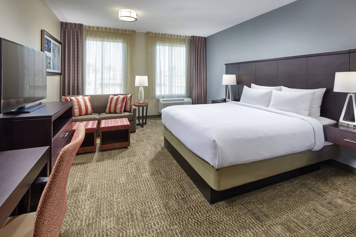 A hotel room with a large bed, a desk with a flat-screen TV, a sofa with red and white cushions, and two side tables with lamps.