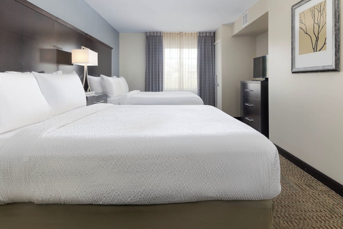 A hotel room with two beds, a desk, a lamp, a TV, and a framed picture on the wall.
