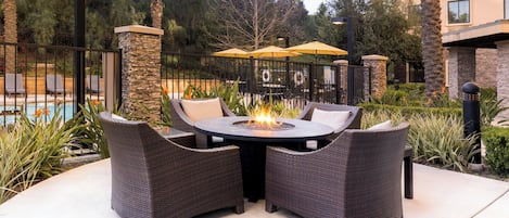 A round outdoor table with chairs, a fire pit, and a pool in the background.