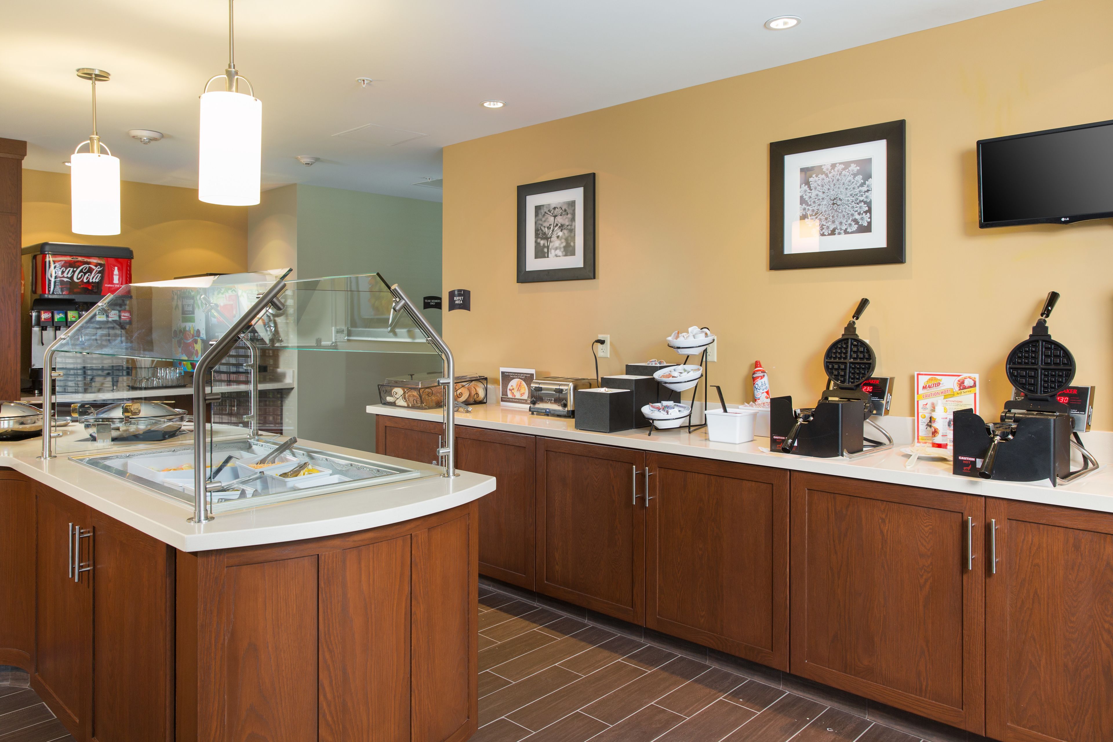 A breakfast bar with a glass enclosure, a Coca-Cola cooler, waffle makers, and a flat-screen TV on the wall.