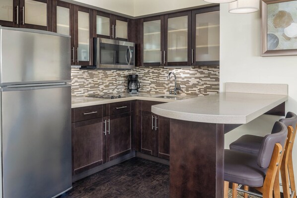 A kitchen with dark wood cabinets, stainless steel appliances, a tiled backsplash, and a bar area with chairs. - Staybridge Suites Corona South by IHG (Corona)